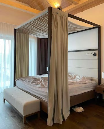 A bedroom with a four poster bed, white linens, and curtains, a cream bench at the foot, and a wooden ceiling.