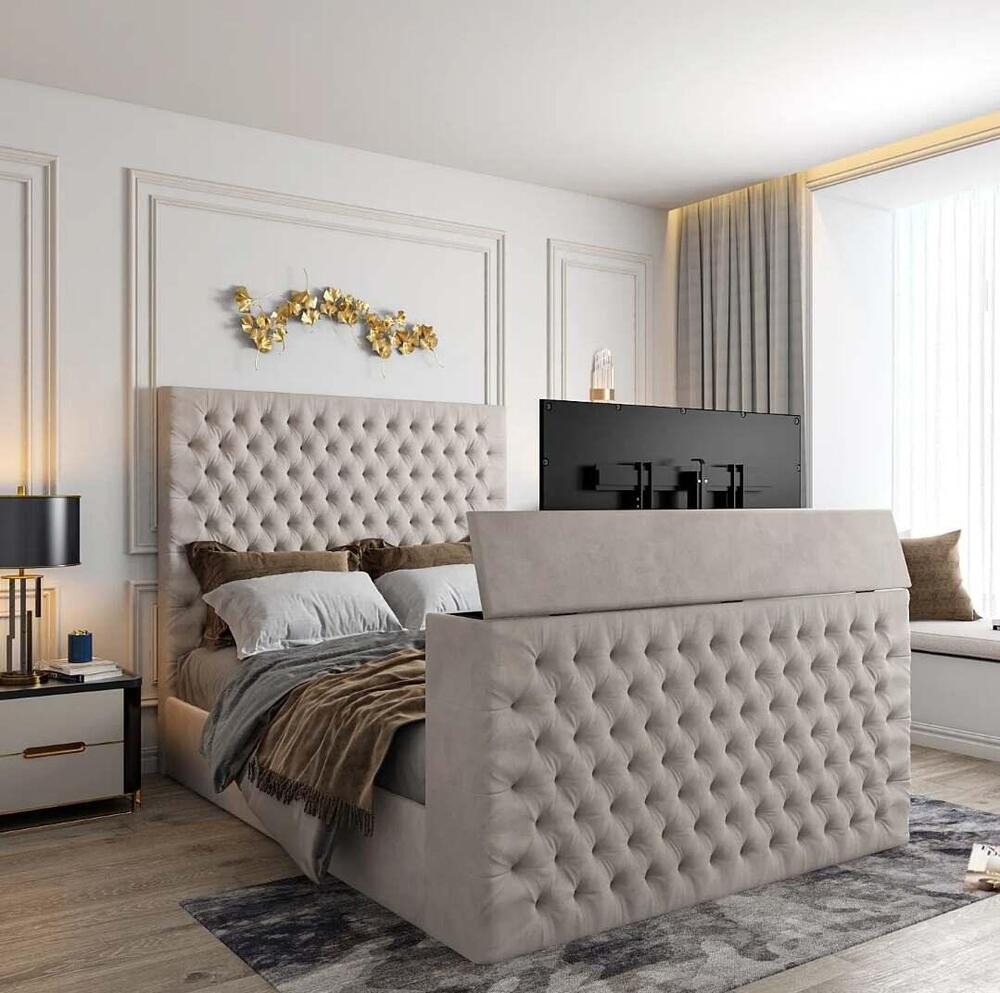 The image features a modern bedroom with a Florence TV bed, showcasing a tall, tufted light gray headboard and footboard. The bed is dressed with gray and brown pillows and a cozy brown throw. A patterned area rug complements the decor. A sleek nightstand holds a black lamp, and above the headboard, abstract gold leaf wall art adds elegance. Tall curtains partially obscure a large window, allowing soft natural light, while a window seat on the right is adorned with plush pillows.