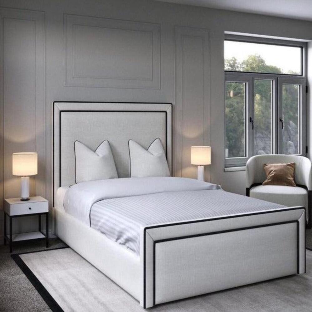 A sleek modern bedroom showcases a linear bed with a white upholstered headboard and black trim. It features white linens, decorative pillows, and matching bedside tables with lamps. A white armchair with a brown pillow adds warmth, while lightly paneled walls and a large window let in natural light. A light gray rug bordered in black completes the look.

