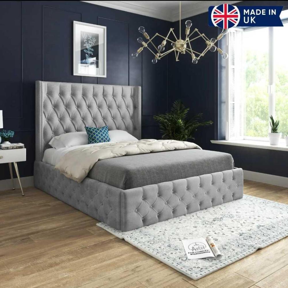 The image displays a modern bedroom with the Short Wing Imperial Bed, featuring a tufted grey upholstered headboard. The bed is dressed in grey bedding with a blue throw pillow. A white nightstand holds a decorative bowl and books. The dark navy wall has a floral picture, and a geometric chandelier hangs above. A light rug extends under the bed, illuminated by natural light from a window near a green potted plant. The UK flag in the corner reads "Made in UK."