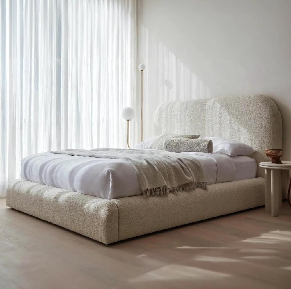 The image shows a serene, minimalist bedroom with a Teddy Boucle Bed featuring a cream upholstered frame and headboard. The bed is adorned with white bedding, a beige throw, and a matching bolster pillow. Sunlight filters through sheer curtains, softly illuminating the room, while a small wooden side table and a modern gold floor lamp add elegant touches to the space.